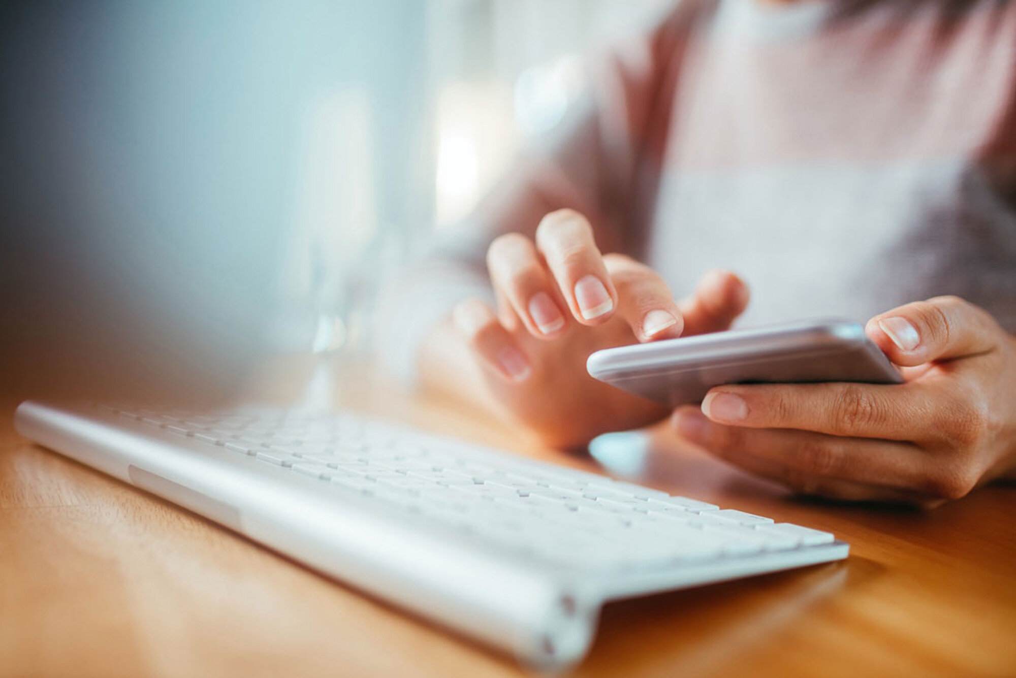 Eine Person hält ein Handy in der Hand. Davor liegt eine Tastatur auf einem Schreibtisch.